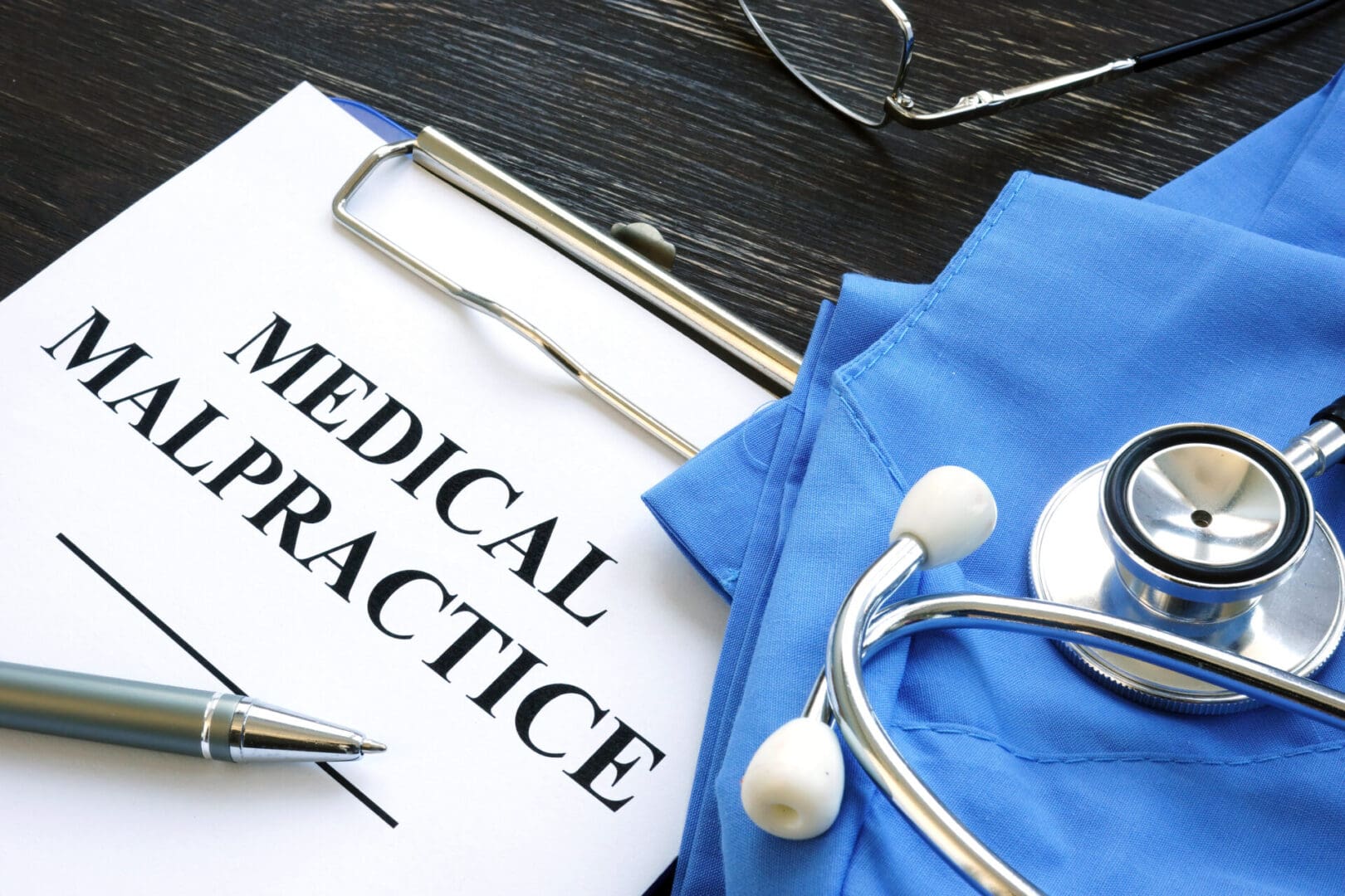 Clipboard with "Medical Malpractice" paper, a pen, stethoscope, and eyeglasses resting on blue scrubs—essential tools for those offering medical record chronology services.