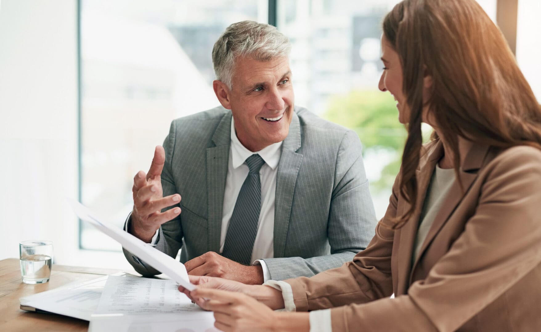 A man and woman in business attire discuss documents, potentially related to medical record chronology services, in a sleek office with a cityscape view.