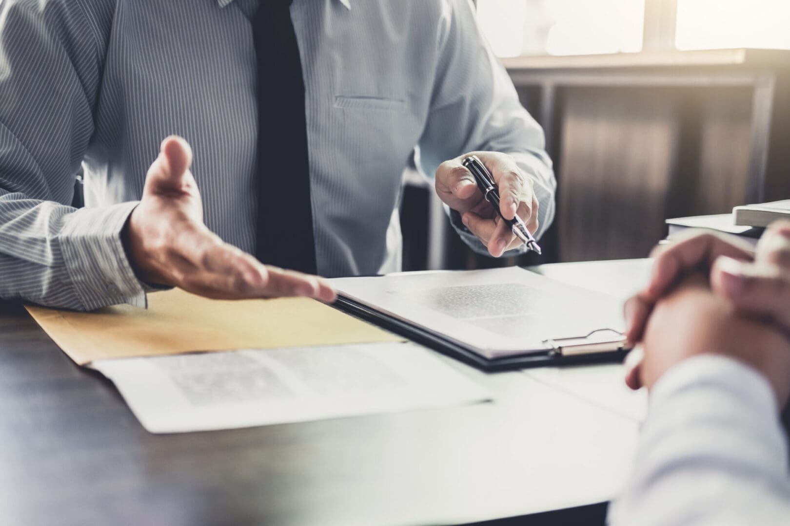 Two people sit at a table, discussing documents. One gestures with a pen over a clipboard while the other clasps their hands, deep in conversation about medical record chronology services.