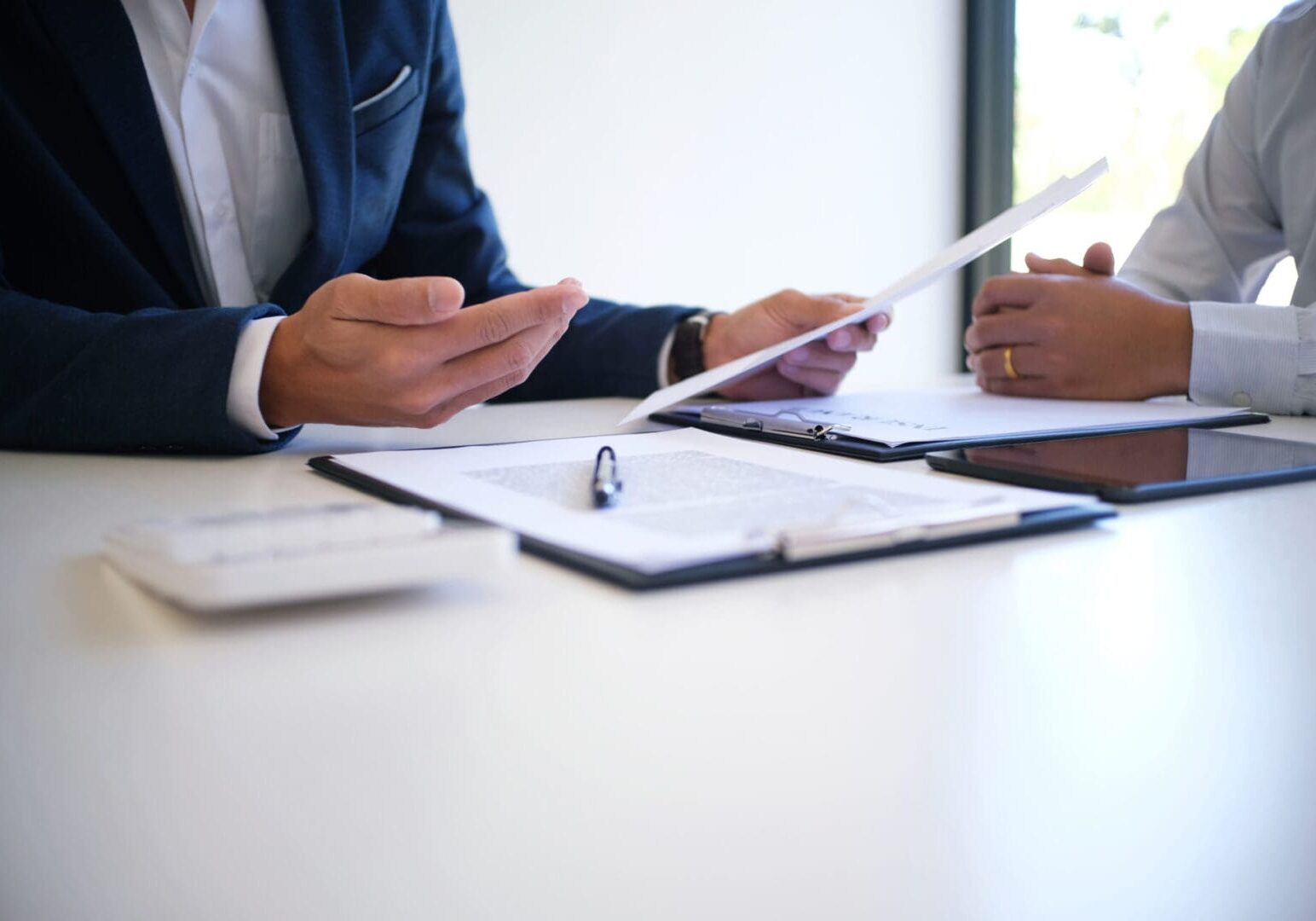 Sales manager giving advice application form document, considering mortgage loan offer for car and house insurance. Two individuals seated at a table discuss documents. One person gestures while the other holds papers, likely reviewing details related to medical record chronology services. A tablet and open binder are on the table, aiding their focused exchange.